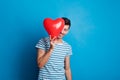 Portrait of a young man in a studio on a blue background, hiding behind red heart. Royalty Free Stock Photo