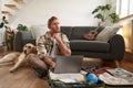 Portrait of young man, traveller going on holiday with a dog, packing his suitcase, sitting with laptop on his luggage Royalty Free Stock Photo