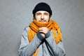 Portrait of young man who feels cold, wearing orange scarf and black beanie hat, on gray background. Royalty Free Stock Photo