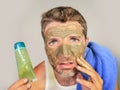 Portrait of young messy funny man in bathroom mirror with green face holding cream male beauty product applying facial mask feelin