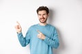 Portrait of young modern man in blue sweatshirt pointing fingers at upper left corner and looking away, smiling pleased Royalty Free Stock Photo