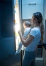 Portrait of young mother holding her baby opens refrigerator to find some food at night Royalty Free Stock Photo