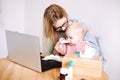 Portrait of young mother sitting at table near laptop, holding little baby, trying to instill medicine into childs nose.