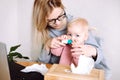 Portrait of young mother sitting at table near laptop, holding little baby, trying to spray medicine into childs nose.