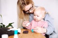 Portrait of young mother woman sitting at table, holding little baby girl, showing how to instill medicine into nose.