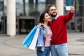 Portrait of young multiracial couple making smartphone selfie, holding bright gift bags after shopping at city mall Royalty Free Stock Photo