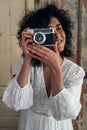 Portrait of young multiracial woman holding vintage style camera taking a picture looking at camera. Vertical image. Royalty Free Stock Photo