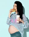 Young pregnant brunette woman in blue jeans, stripy t-shirt eats bites fresh hamburger holds her drink