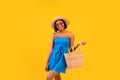 Portrait of young pretty black lady in straw hat and blue dress smiling, holding trendy beach bag over orange background Royalty Free Stock Photo
