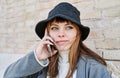 Portrait young pretty Caucasian girl, dressed in a gray jacket, a white sweater and a hat, in the city, talking on the mobile phon Royalty Free Stock Photo