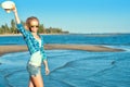 Portrait of young pretty smiling slim blond wearing mirrored heart shaped sunglasses walking along the seaside and waving a hat