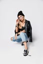 Portrait of young punk style blonde girl in torn jeans, leather top, leather jacket and chrome boots sitting squatting Royalty Free Stock Photo