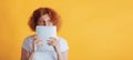 Portrait of young red-haired woman covering face with tablet, thinking, poisng isolated over yellow studio background Royalty Free Stock Photo