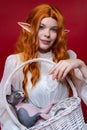 Portrait of young redhead woman cosplayer elf with wicker basket in hands on red background