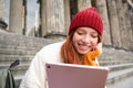 Portrait of young redhead woman sitting outdoors on stairs, reading e-book on digital tablet, wearing red hat and warm Royalty Free Stock Photo