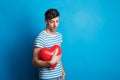 Portrait of a sad young man in a studio on a blue background, holding red heart. Royalty Free Stock Photo