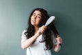 Portrait of a young sad woman trying to comb her long dark tangled hair with hairbrush. Blue background. The concept of Royalty Free Stock Photo