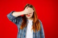 Portrait of young scared woman covering eyes with one hand while standing against red background. Confused girl close