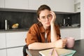 Portrait of young serious woman, working from home in glasses, sitting with folder and documents in kitchen, looking Royalty Free Stock Photo