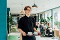 Portrait of young smiling affable waiter carrying on tray with coffee cups to client table in cafe. Hospitality service Royalty Free Stock Photo