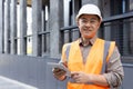 Portrait of a young smiling Asian man standing near a building in a hard hat and vest, holding a lancet and looking at Royalty Free Stock Photo