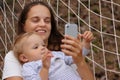 Portrait of young smiling attractive woman with her daughter, mother and child relaxing in hammock in forest and using smart phone Royalty Free Stock Photo