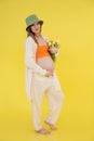 Portrait of young smiling barefoot pregnant woman wear green bucket hat, orange top, standing, holding bunch of tulips. Royalty Free Stock Photo