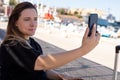 Portrait of young smiling beauteous woman sitting near beach embankment, taking selfie in shadow, posing on sunny day. Royalty Free Stock Photo