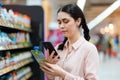 Portrait of young smiling beautiful caucasian woman scan qr code of product using smartphone. Aisle and shelves in