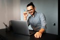 Portrait of young smiling confident man working home at laptop, holding pencil in hand, adjusts his glasses, wearing eyeglasses Royalty Free Stock Photo