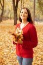 Portrait of young smiling flawless woman standing among yellow fallen leaves in forest holding bunch of leaves, posing.