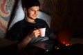 Portrait of young smiling guy working home at laptop in night room. Lying on bed against pillow, holding pink coffee mug. Royalty Free Stock Photo