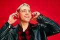 Portrait of young smiling handsome man with blonde hair, in leather jacket listening to music in headphones against red Royalty Free Stock Photo