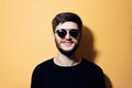 Portrait of young smiling hipster, dressed in black, wearing sunglasses, isolated on yellow background. Royalty Free Stock Photo