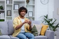 Portrait of young smiling hispanic man at home on sofa smiling and looking at camera, holding phone in hands using app Royalty Free Stock Photo