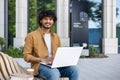 Portrait of a young smiling Indian man sitting on a bench outside with a laptop on his lap, working and studying online Royalty Free Stock Photo