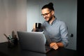 Portrait of young smiling man with hair bun working home at laptop, drinking coffee from black cup, wearing eyeglasses and shirt, Royalty Free Stock Photo