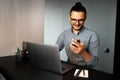 Portrait of young smiling man working home at laptop, looks at the smartphone screen, wearing eyeglasses and shirt. Royalty Free Stock Photo