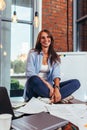 Portrait of young smiling student sitting on table covered in papers in lotus pose preparing for exam in study room