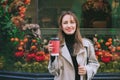 Portrait of young smiling woman in coat enjoying drink in reusable coffee cup walking on street with shop windows
