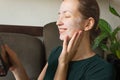 Portrait of young smiling woman with a cosmetic mask on her face. Looking at herself in a hand mirror. Royalty Free Stock Photo