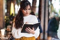 Portrait of young smiling woman standing in cafe near window, writing in notebook. Girl writes notes diary, makes business plan Royalty Free Stock Photo