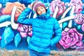 Portrait of young smiling woman wearing in blue down jacket and knitted warm hat posing against wall painted with