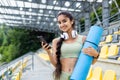 Portrait of young sporty Indian woman standing on stadium tribune in suit and headphones, holding exercise mat and phone Royalty Free Stock Photo