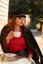 portrait of a young stylish beautiful woman sitting in an outdoor cafe in a red sweater and black hat, street style Royalty Free Stock Photo