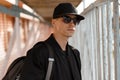 Portrait of a young stylish man hipster in stylish sunglasses in a black cap in a fashionable jacket with a backpack on his Royalty Free Stock Photo