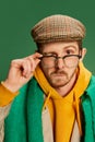 Portrait of young stylish man in trendy clothes, glasses, cap and scarf posing isolated over green background. Royalty Free Stock Photo