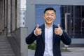 Portrait of a young successful Asian male businessman standing outside an office building in a suit, smiling, looking at Royalty Free Stock Photo