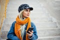 Portrait of young teenage blonde girl with smartphone in hand, wearing blue denim jacket with orange scarf and plaid cap. Royalty Free Stock Photo