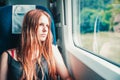 Young teenager redhead girl with long hair looks out the window while sitting in the train.